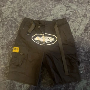 Svarta shorts från Corteiz - Snygga svarta shorts från Corteiz med praktiska fickor och ett coolt tryck framtill. De har en justerbar snörning i midjan och en gul etikett på sidan. Perfekta för en avslappnad stil. Aldrig använd.