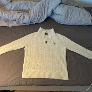 Vit stickad tröja från Polo Ralph Lauren - Säljer en stilren vit stickad tröja från Polo Ralph Lauren med kabelstickat mönster och en broderad logotyp på bröstet. Tröjan har en hög krage med dragkedja och långa ärmar. 
