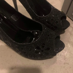 Svarta glittriga pumps med öppen tå - Snygga svarta pumps med glittrig yta och öppen tå. Skorna har ett dekorativt blommönster och är perfekta för en festlig kväll. Klacken ger en elegant touch och de är bekväma att bära.