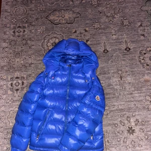 Moncler Maya  - Säljer en snygg blå dunjacka från Moncler. Jackan har en hög krage och en praktisk huva. Den är utrustad med dragkedja och har Monclers logotyp på ärmen. Perfekt för kyliga dagar! Jag säljer den då den är för liten och inte kommer till användning! Skicket är 9 av 10 använd ett fåtal gånger. Priset är inte hugget i sten! Skriv vid funderingar