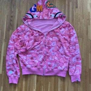 Rosa camo hoodie från A Bathing Ape - Säljer en cool rosa camo hoodie från A Bathing Ape med dragkedja och broderade detaljer på huvan. Perfekt för dig som vill sticka ut med en unik stil. Hoodien har långa ärmar och en bekväm passform.