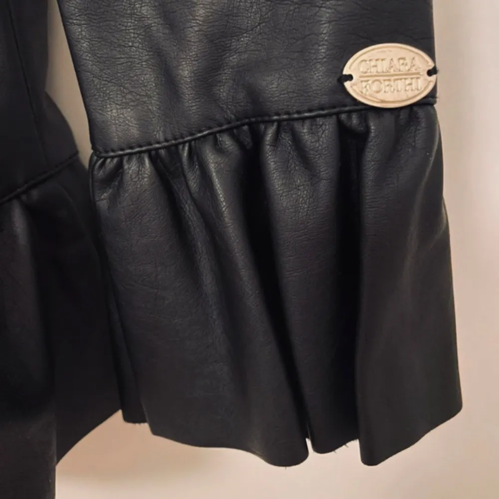 Snygg svart jacka från Chiara Forthi i skinnimitation med dragkedja framtill. Jackan har volangdetaljer vid ärmslut och nederkant, vilket ger en trendig look. Perfekt för att lyfta din outfit!. Takit.