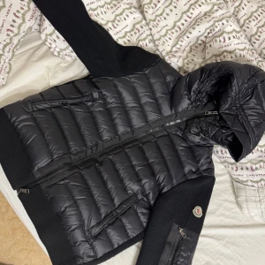 Svart jacka från Moncler - Säljer en snygg svart jacka från Moncler med quiltad design och dragkedja. Jackan har långa ärmar och en huva för extra skydd. Perfekt för kyligare dagar. Cardigan. 