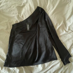 Svart oneshoulder-top från Boohoo - Helt ny!! Snygg svart oneshoulder-top från Boohoo i ett glansigt material. Perfekt för en stilren look med en lång ärm och asymmetrisk design. 