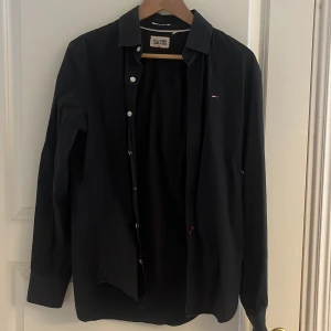 Svart hilfiger skjorta, slim fit i storlek M. Skick 8/10. Väldigt fint skick och inga fläckar eller hål på skjortan/ skriv dm gällande frågor eller vid intresse! - Snygg svart skjorta från Hilfiger Denim med klassisk krage och knappar framtill. Perfekt för en stilren look. Den har långa ärmar och en liten logotypdetalj på bröstet.