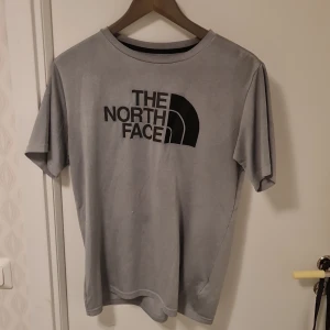 Grå t-shirt från The North Face - Säljer en grå t-shirt från The North Face med deras ikoniska logga tryckt i svart på bröstet. T-shirten har en klassisk rund hals och korta ärmar, perfekt för en avslappnad stil.
