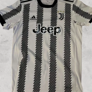 Juventus fotbollströja  - Snygg vit och svart fotbollströja från Adidas med ett unikt triangulärt mönster. Tröjan har korta ärmar och klubbens logga på bröstet. Perfekt för fotbollsfans som vill sticka ut på planen eller läktaren.