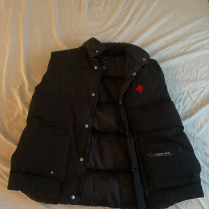 Svart dunväst från Canada Goose - Säljer en svart dunväst från Canada Goose med en röd logotyp på bröstet. Västen har knappar och dragkedja framtill samt två stora fickor
