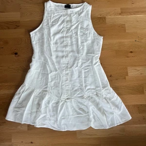 Vit ärmlös klänning från ASOS Design - Säljer en söt vit ärmlös klänning från ASOS Design. Klänningen har knappar framtill och en volangdetalj vid fållen. Perfekt för varma sommardagar eller som studentklänning! Klänningen är helt ny med prislappen kvar och i ett superskönt material! 