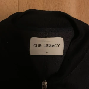 Mörkblå/Svart jacka med ränder från Our Legacy - Mörkblå / Svart jacka från Our Legacy med vita ränder längs ärmarna. Jackan har dubbeldragkedja framtill och är tillverkad i en blandning av polyester och bomull. Perfekt för en stilren look.