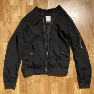  Svart bomberjacka  - Snygg svart bomberjacka från Premium med dragkedja framtill och fickor med knappar. Jackan har en klassisk bomberstil med ribbade muddar vid ärmslut och nederkant. Perfekt för en stilren look.