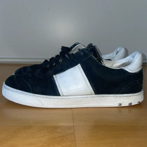 Valentino Garavani sneakers i svart och vitt - Snygga sneakers från Valentino Garavani i svart mocka med vita detaljer. Skorna har en rund tå och snörning framtill. Sulan har ett coolt kamouflagemönster och små nitar runt hälen för extra stilpoäng.