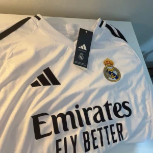 Real Madrid hemmatröja 2023/24 från Adidas - Säljer en vit Real Madrid hemmatröja från Adidas med svarta detaljer och klubbmärke på bröstet. Tröjan har korta ärmar, klassisk V-ringning och 'Emirates Fly Better' tryckt på framsidan. Perfekt för alla Real-fans!