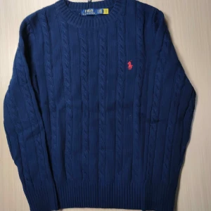 Mörkblå stickad tröja från Polo Ralph Lauren - Säljer en klassisk mörkblå stickad tröja från Polo Ralph Lauren med rund halsringning och kabelstickat mönster. Tröjan har lång ärm och en röd broderad logga på bröstet. Perfekt för en stilren och tidlös look. Storlek M, vid fundering är det bara att skicka dm