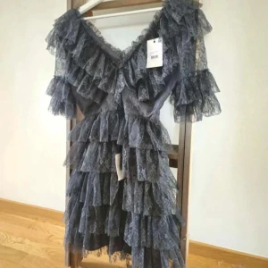 Grå volangklänning i spets från By Malina - Supersnygg grå kortklänning från By Malina med flera lager volanger och spetsdetaljer. Klänningen har v-ringning och är kortärmad, perfekt för dig som vill sticka ut på festen. Tillverkad i polyester med stretchig känsla.