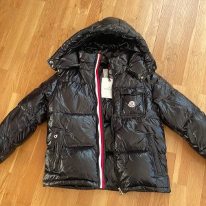 Svart pufferjacka från Moncler - Säljer en snygg svart pufferjacka från Moncler i nytt skick och aldrig andvänd. Jackan har en cool glansig finish. Den är perfekt för vintern med sin varma vaddering och har en praktisk huva. Passar perfekt för kalla dagar! Jag säljer den därför den inte passade mig. Skriv för frågor, prutbar vid snabb affär