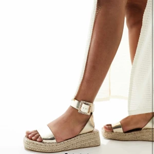 Guldiga sandaler med platåsula - Säljer ett par snygga sandaler i guldigt syntetmaterial med bred rem över tårna och justerbart spänne vid ankeln. Skorna har en flätad platåsula som ger extra höjd och somrig känsla. Perfekta till varma dagar och lätta outfits.