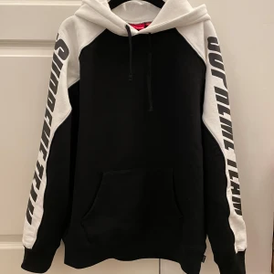 NY Svart och vit hoodie från Supreme - Säljer en svart och vit hoodie från Supreme med stora tryckta bokstäver på ärmarna. Tröjan har huva med dragsko och en stor magficka. Perfekt för dig som gillar streetwear och vill sticka ut.