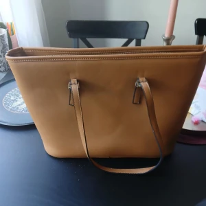 Kamelbrun axelväska från Michael Kors - Säljer en stilren kamelbrun axelväska från Michael Kors med långa handtag och dragkedja upptill. Väskan är rymlig och passar perfekt för dig som vill ha plats med allt du behöver under dagen. Klassisk design med diskret logga framtill.