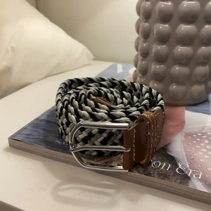 Svart och vit flätat bälte med bruna skinn-detaljer - Snyggt flätat bälte i svart och vitt med bruna skinn-detaljer vid spännet. Bältet har ett silverfärgat dubbelt spänne och passar perfekt till jeans eller chinos. Ger en cool och modern touch till outfiten.