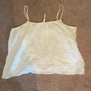Vit linnetopp med tunna axelband - Säljer en vit, luftig linnetopp med tunna axelband och knytning fram. Toppen är ärmlös och har en avslappnad passform, perfekt för varma dagar.