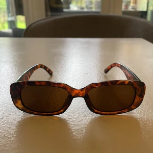 Bruna rektangulära solglasögon med leopardmönster - Snygga solglasögon med rektangulära bågar i brun plast och leopardmönster. Glasen är mörka och bågarna har en trendig, kantig form som ger en cool retrostil.