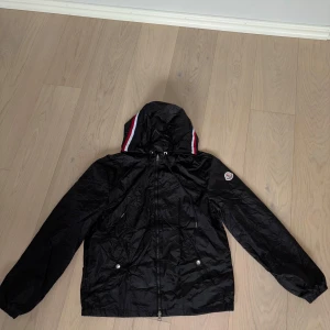 Svart windbreaker från Moncler med huva - Svart windbreaker från Moncler med huva och dragkedja. Jackan har Moncler-logga på ärmen och rödvitblå detalj på huvan. Två fickor framtill med knappar och lätt glansigt material. Aldrig använd utan den var för liten 10/10 skick. Allt scannar.
