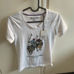 Vit t-shirt från U.S. Polo Assn. - Vit t-shirt från U.S. Polo Assn. med tryck av två polospelare på hästar framtill. Klassisk rund halsringning och korta ärmar. Tillverkad i mjuk bomull, perfekt för en avslappnad stil.