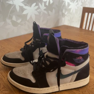 Nike Air Jordan 1 Zoom Paris Saint-Germain - Säljer ett par Nike Air Jordan 1 Zoom Paris Saint-Germain med svart, grå och lila färgskala. Skorna har en skimrande blå Swoosh, PSG-detaljer på hälen och klassisk snörning. Hög modell med rund tå och platt sula. Nypris ligger på runt 5500 för den här storlek 