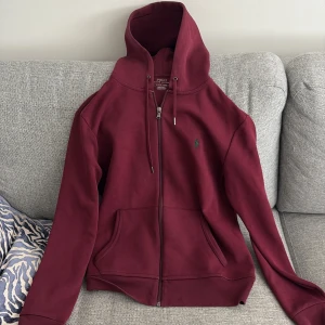 Vinröd hoodie från Polo Ralph Lauren - Säljer en vinröd hoodie från Polo Ralph Lauren med dragkedja och huva. Tröjan har två fickor framtill och en broderad grön logga på bröstet. Perfekt för en avslappnad och stilren look.