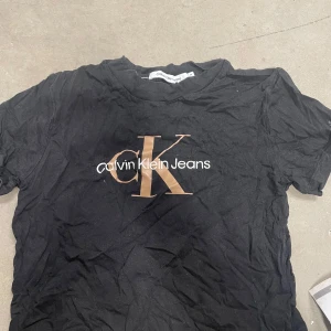 Svart t-shirt från Calvin Klein Jeans - Svart t-shirt från Calvin Klein Jeans med stort CK-tryck i beige och vitt framtill. Klassisk rund halsringning och korta ärmar. Perfekt för en stilren och enkel look.