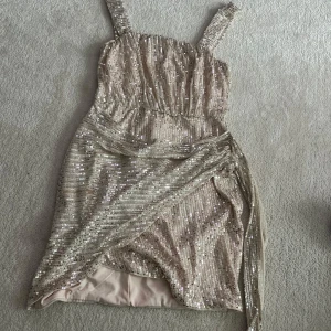 Glittrig kort klänning med omlott och paljetter - Säljer en glittrig kort klänning med breda axelband och omlott-detalj framtill. Klänningen är täckt av paljetter i en ljus champagnefärg och har en markerad midja. Perfekt för festliga tillfällen!