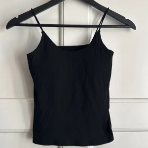 Svart linnetopp med smala axelband - Enkel och stilren svart linnetopp med smala axelband. Toppen är tillverkad i 100% polyester och har en rak passform. Perfekt att matcha med jeans eller kjol för en clean look.