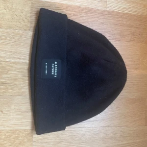 Svart mössa från Alternate Future - Svart mössa med uppvikt kant och liten vävd logotyp framtill från Alternate Future. Enkel och stilren design som passar till det mesta. Perfekt för kyliga dagar.