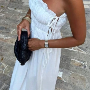 Vit axelbandslös långklänning med knyt - Säljer en superfin vit axelbandslös långklänning med smock upptill och dekorativa knytningar på sidan. Klänningen har en luftig och flowy passform, perfekt för varma dagar. Stilren och enkel design som passar till många tillfällen. Säljer även en svart!