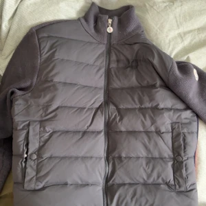 Grå dunväst från Moncler med stickade ärmar - Säljer en grå dunväst från Moncler med stickade ärmar och hög krage. Västen har dragkedja framtill, två fickor med knappar och Moncler-logga längs sidorna i rött. Perfekt för lager-på-lager och snygg streetstil.