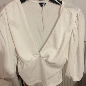 Vit v-ringad blus från Gina Tricot - Säljer en vit blus från Gina Tricot med djup v-ringning och långa ärmar. Blusen har lätt puff vid axlarna och är tillverkad i ett strukturerat tyg som ger en snygg och modern look.
