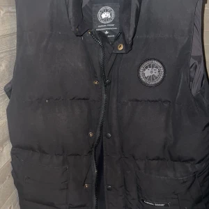 Canada Goose väst black label (billigare vid snabbare affär) - Säljer en svart dunväst från Canada Goose med klassisk logotyp på bröstet. Västen har stora fickor framtill, dragkedja och knappar. Perfekt för lager-på-lager och håller dig varm under kyliga dagar. Äkta goose väst med tillbehör som johanells påse och klädhängare med följer billigare vid snabbare affär