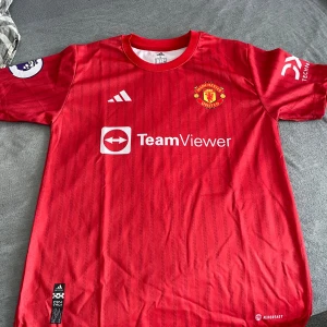 Manchester United fotbollströja Ronaldo 7 - Säljer en röd Manchester United matchtröja med Ronaldo 7 på ryggen. Tröjan har vita detaljer, klubbmärke på bröstet och TeamViewer sponsortryck. Klassisk design med korta ärmar och Adidas-logga. Perfekt för fans!