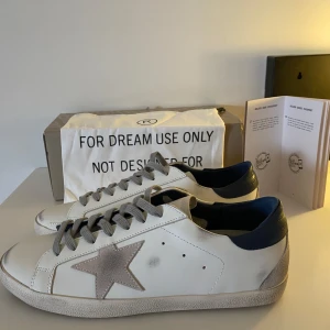 Golden Goose skor - Snygga vita sneakers från Golden Goose med grå stjärna på sidan och mörkblå häl. Skorna har grå snörning och en platt sula. Perfekta för dig som gillar en trendig och avslappnad stil. Ej äkta. Allt org till kommer, alltså påsen, box och manualen. Skriv vid minsta fundering 