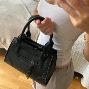 Svart väska - Svart väska med detaljer och dragkedja på ena sidan. Köpt på ginatricot. Små skavanker på några få ställen. En längre axelrem kommer med!😘💕