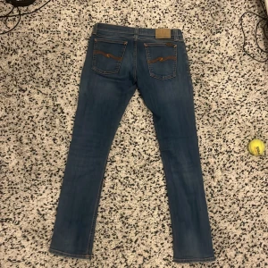 Blå jeans från Nudie Jeans - Snygga blå jeans från Nudie Jeans med klassisk femficksdesign och kontrastsömmar på bakfickorna. Perfekta för en avslappnad stil med en modern touch. Midjan är normalhög och passformen är slim fit. Använda få gånger nästan helt nya 