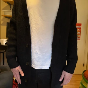 Svart stickad kofta med knappar från cos  - Säljer en svart stickad kofta med v-ringning och stora knappar framtill. Koftan har lång ärm och en klassisk, lite lös passform. Perfekt att slänga på sig över en t-shirt eller skjorta. 