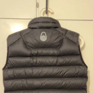 Black dunväst från Sail Racing - Snygg black dunväst från Sail Racing med hög krage och dragkedja framtill. Västen har två fickor med dragkedja och en diskret logga på bröstet. Perfekt för lager-på-lager under kyliga dagar. Köptes för 1200kr nypris. Säljs för 600kr