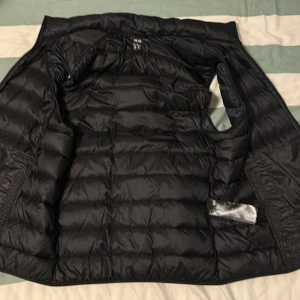 Svart dunväst från Uniqlo - Säljer en svart dunväst från Uniqlo med hög krage och diskret design. Västen är lätt och smidig, perfekt som extra lager under kyliga dagar. Stängs med dragkedja framtill och har en quiltad yta. Den här på någon plats litet hål där det kan komma ut lite bomull, de är så lite så att jag inte ens hittade stället. Storlek M. (Uppmöte)