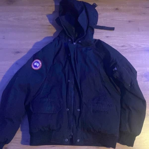 Svart dunjacka med huva från Canada Goose - Säljer en svart dunjacka från Canada Goose med huva och klassisk logotyp på ärmen. Jackan har dragkedja framtill, stora fickor och ribbade muddar. Perfekt för kalla dagar och riktigt snygg streetstil. Pm för frågor, pris ej hugget i sten