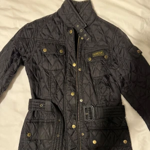 Svart jacka från Barbour - Säljer en svart quiltad jacka från Barbour International med bälte i midjan och guldiga knappar. Jackan har hög krage, fyra fickor framtill och Barbour-logga på bröstet.  I väldigt bra skick, har dock smink fläck vid krage går att få bort i tvätt.  Inga repor eller slitage endast fläckar som går tar bort i tvätt, se bilder.