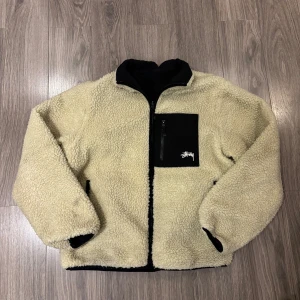 Stussy Fleece - Säljer en fet reversibel teddyjacka från Stussy. Ena sidan är beige teddy med svart bröstficka och stor 8-ball på ryggen, andra sidan är svart med beige teddykrage och Stussy-logga på bröstet. Dragkedja framtill och fickor på båda sidor.