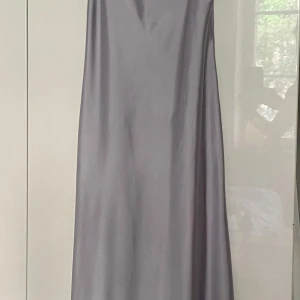 Grå långklänning från Zara - Säljer en elegant silverfärgad långklänning från Zara i satin med smala axelband och v-ringad urringning. Klänningen har en öppen rygg med snygg detalj och en mjukt följsam passform. Perfekt för speciella tillfällen. Använd en gång till fest. En liten defekt orsakad av för varm strykning finns (se sista bild), men inget som stör. I övrigt fint skick. Storlek S