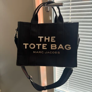 Marc Jacobs the tote bag väska svart - Säljer en svart tote bag från Marc Jacobs med tydlig vit text på framsidan. Väskan har både handtag och en justerbar axelrem, perfekt för att bära på olika sätt. Tillverkad i kraftig canvas och rymmer det mesta du behöver till vardagen.